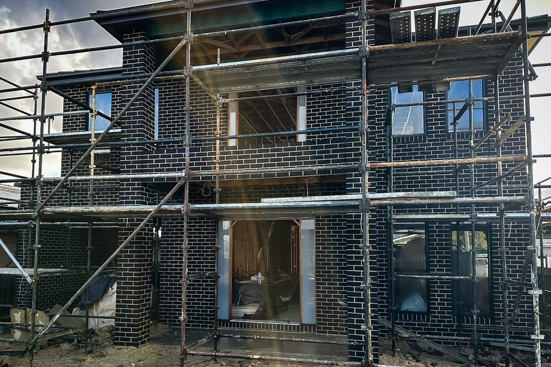 Two-storey home in blue-black face brick under construction behind scaffolding